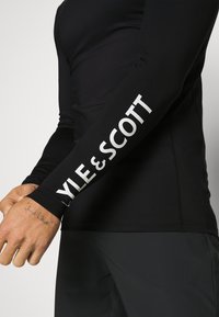 Long-sleeve black shirt made of stretch fabric, featuring a white "KYLE & SCOTT" logo along the left arm and flatlock stitching details.