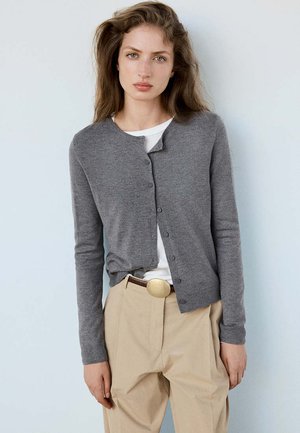 Jeune femme aux longs cheveux portant un cardigan gris boutonné sur une chemise blanche et un pantalon beige avec une grande boucle de ceinture.
