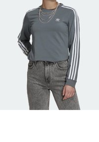 Långärmad grå skjorta med vit tre-stripesdesign på ärmarna och liten logotyp på bröstet, matchad med högmidjade grå denimjeans.