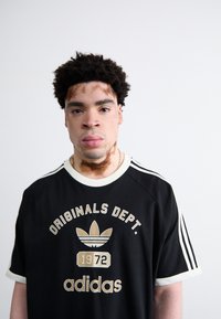 Melnā Adidas t-krekls ar baltu apdari, kurā ir zelta grafiskais teksts "ORIGINALS DEPT. 1972" un logo. Īsas piedurknes un trīs baltas svītras uz pleciem.