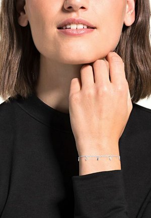Bracciale in argento con piccoli charm ovali appesi. La mano della modella poggia sul mento, indossando un top nero con scollatura alta.