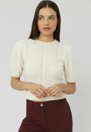 Pull en tricot crème avec des manches bouffantes courtes, un motif en diamants texturé et un ourlet côtelé. Porté avec un pantalon ajusté bordeaux.