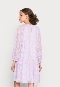 Vestido com padrão floral de lavanda, mangas longas transparentes, cintura franzida e detalhe em forma de lágrima nas costas. Textura suave e silhueta divertida.