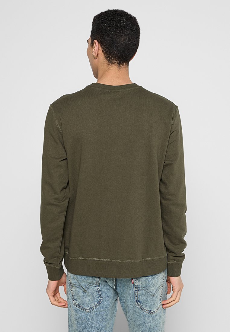 Olivgrüner Pullover, Rundhalsausschnitt, lange Ärmel, gerippte Bündchen und Saum, Baumwollmischung, glatte Textur. Getragen mit hellblauen Jeans.