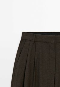 Pantalon plissé marron foncé taille haute avec passants de ceinture, partiellement visible sur fond blanc.