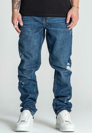 Blå denim jeans med slim fit, der har slidte detaljer og en medium vask. Kombinationen er med hvide sneakers på en grå baggrund.