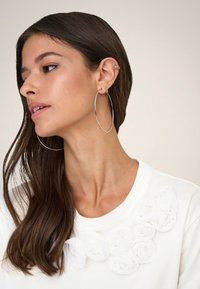 Jeune femme aux longs cheveux bruns portant de grandes boucles d'oreilles argentées en anneau et un haut blanc décoré de fleurs en tissu blanc.
