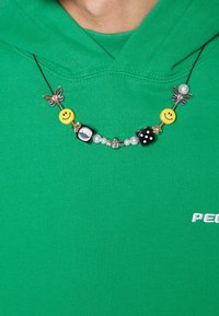 Collana di perline colorate con faccine sorridenti gialle, dadi bianco e nero, perline trasparenti e accenti metallici a forma di farfalla, indossata sopra un felpa verde.