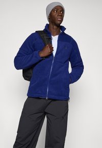 Chaqueta de forro polar en azul profundo, con cremallera frontal, cuello alto y bolsillos laterales, combinada con un gorro gris y una mochila negra.