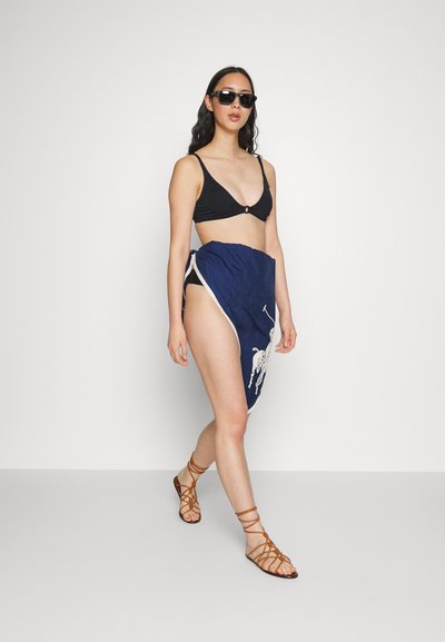 Biquíni preto com padrão em ribana e um detalhe de hardware circular, combinado com um lenço azul com um design de jogador de pólo branco e sandálias castanhas de tiras.