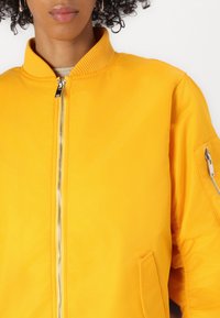 Giacca bomber gialla con colletto a costine, chiusura con zip e tasche laterali. Caratterizzata da una texture liscia e maniche corte.