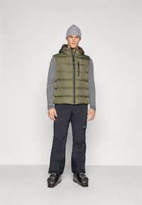 Doudoune sans manches pour homme en vert olive, avec une chemise à manches longues grise, un pantalon noir et des bottes de ski noires. Comprend une poche zippée et une capuche avec des accents.