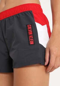 Calvin Klein Badehose in Grau, Rot und Weiß. Elastischer Bund, seitliches Design und auffälliges rotes Logo auf dem linken Bein.