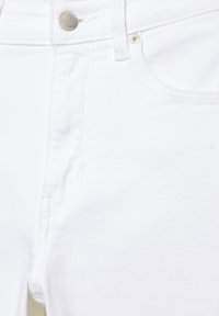 Bílé džíny z denimu, detailní záběr ukazující přední kapsu, poutko na opasek a stříbrný kovový knoflík se šitími.