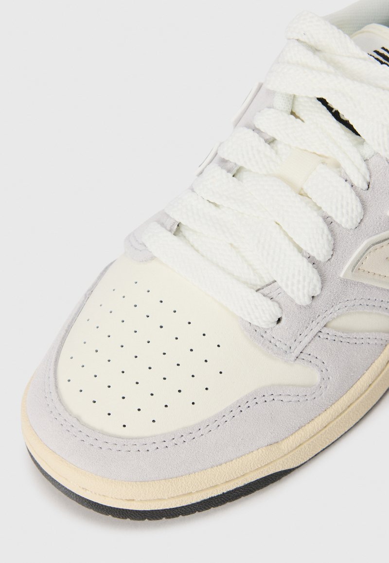 Sneaker in suede grigio chiaro con punta in pelle bianca perforata, stringhe piatte bianche e suola in gomma testurizzata. Presenta un design liscio e arrotondato.