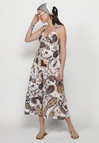 Balta maksi suknelė su spalvingais paisley raštais, turinti V formos iškirptę, plačias petnešas ir sklindančią sijoną. Modelis derina ją su juoda ir balta galvos juosta.