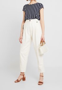 Maglia a righe blu navy e bianca a maniche corte, pantaloni a vita alta color crema, sandali con tacco a blocco marroni e una borsa trasparente con manici in legno.