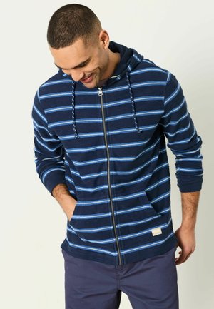 Mann trägt eine marineblaue Kapuzenjacke mit hellblauen horizontalen Streifen und hat die Hände in den Vorder taschen, schaut nach unten und lächelt.