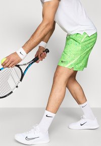 Πράσινα σορτς τένις με βούλες, λευκό αθλητικό μπλουζάκι και λευκά αθλητικά παπούτσια με μαύρες λεπτομέρειες. Ο παίκτης κρατά μια μπάλα του τένις και ρακέτα.