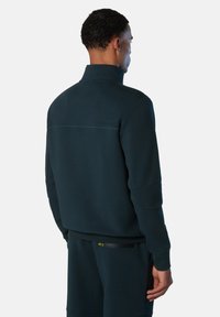Sudadera de color azul petróleo oscuro con cuello alto, tela texturizada y un logo sutil en la parte posterior. Incluye un detalle de bolsillo con cremallera en los pantalones.