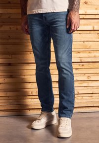 Jasnoniebieskie dżinsy o prostym kroju, z delikatnym efektom przetarć i kontrastowym przeszyciem, w połączeniu z beżowymi sneakersami i neutralnym t-shirtem.