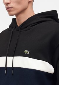Gros plan d'une personne portant un sweat à capuche noir, blanc et marine avec un logo crocodile vert sur la poitrine et des cordons noirs à la capuche.