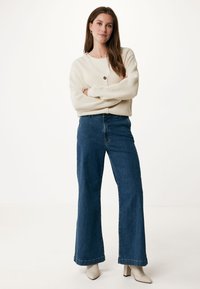 Krémový žebrovaný cardigan s detaily knoflíků, nošený přes béžový top, spárovaný s vysokými modrými širokými džínami a krémovými botami na podpatku.