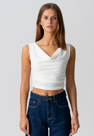 Noorpruunide sirgete juuste ja ilma varrukateta valget lühikest topi ning kõrge vöökohaga tumesiniseid teksaseid kandev noor naine seisab ühevärvilise halli tausta ees.