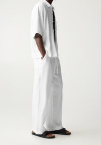 Homme portant un pantalon en lin blanc ample et une chemise à manches courtes assortie, avec des sandales noires, debout devant un fond clair uni.