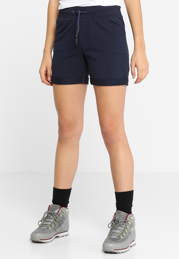 Jambes portant un short bleu marine à cordon, des chaussettes noires aux chevilles et des chaussures de randonnée grises, debout sur une surface blanche.