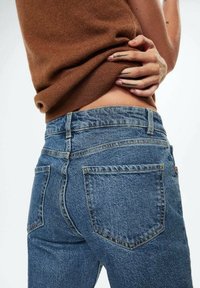 Jean en denim bleu taille haute avec un design classique à cinq poches, présentant un léger délavage, des détails de couture et une coupe droite.