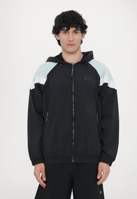 Črna lahka jakna s kapuco, bele in svetlo modre akcenti na ramenih, sprednji zadrgo in stranskimi žepi. Gladka tekstura tkanine.