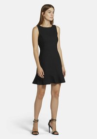 Schwarzes ärmelloses Kleid mit einem taillierten Oberteil, ausgestelltem Saum und gerippter Textur. Kombiniert mit schwarzen, filigranen High-Heels.