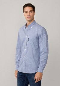 Camisa de botones de patrón a cuadros en azul y blanco, hecha de algodón. Presenta mangas largas y un bolsillo en el pecho, combinada con jeans oscuros.