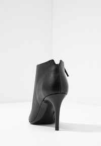 Stivaletto in pelle nera alla caviglia con punta affilata, alto tacco a stiletto e dettaglio con zip posteriore; texture liscia e design minimalista.