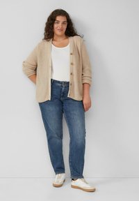 Beige gebreide cardigan met knopen, gelegd over een wit t-shirt. Gepaard met rechte blauwe jeans en witte sneakers met rubberen zolen.