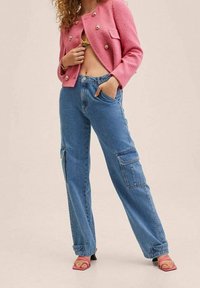 Blazer rosa tejido con botones dorados, combinado con jeans cargo azul claro con bolsillos laterales y corte relajado. Sandalias de tacón alto rosas.