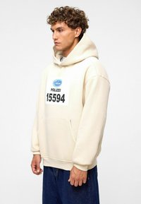Beige hoodie met een voorzak, met een blauw logo en zwarte tekst "POLIZEI 15594" op de borst. Gemaakt van een katoenmix.