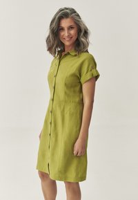 Abito camicia in lino verde con colletto, maniche corte arrotolate e chiusura con bottoni. Presenta ricami decorativi sul lato inferiore.