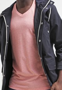 Czarna wodoodporna kurtka z kapturem, z metalowymi akcentami i kieszenią z przodu, noszona na jasnoróżowej koszulce z dekoltem w serek o miękkiej fakturze.