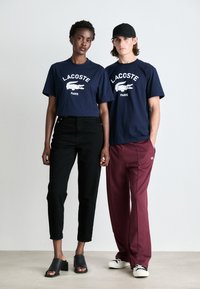 Tricouri Lacoste bleumarin cu logo alb, pantaloni negri cu talie înaltă, pantaloni de sport vișiniu și mules negri cu toc deschis. Design minimalist.