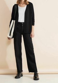 Svart cardigan över en vit t-shirt, kombinerad med högmidjade svarta raka jeans, med fransade kanter och svarta snörskor.