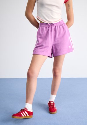 Purple shorts with white side stripes and elastic waistband, paired with red sneakers and white socks, set against a light blue background.
