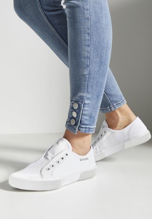 White canvas sneakers with rubber soles, round toe, and lace-less design. Worn with light blue jeans featuring button accents at the hem.
