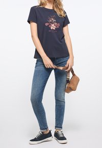 Kvinna klädd i marinblå blommig t-shirt, blå jeans, marinblå sneakers, som håller en brun läderaxelväska i remmen.