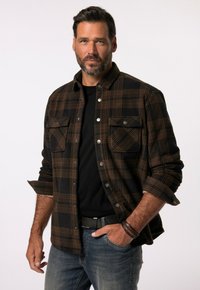 Camisa de franela a cuadros marrón y negra con dos bolsillos en el pecho, botones de metal y mangas arremangadas, llevada sobre una camiseta negra. Pantalones vaqueros visibles.