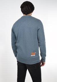 Ljusblå sweatshirt i mjukt tyg med långa ärmar och rund halsringning, med orange logotyp och text på nedre ryggen.