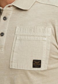 Polo beige à manches courtes avec un col texturé, deux boutons et une poche avant avec un patch noir.
