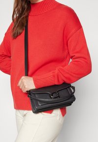 Zwarte leren crossbodytas met een textuurrijke afwerking en een ontwerp met een gebogen klep, vastgehouden tegen een rood gebreide trui.