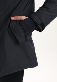 Mørkeblå jakke med en struktureret overflade, der har en lynlåslomme og velcro-justering ved ærmerne, som fremhæver et strømlinet, moderne design.
